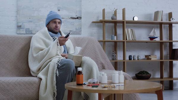 sick man in warm beanie and blanket clicking tv channels near medicaments on table