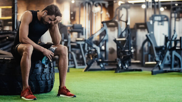 tired sportsman in earphone sitting on car tire and holding sports bottle