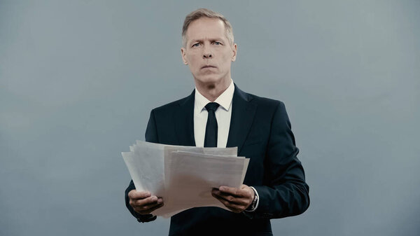 Mature manager in suit holding documents and looking at camera isolated on grey 