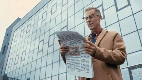 Low angle view of businessman in earphone and coat reading newspaper on urban street 