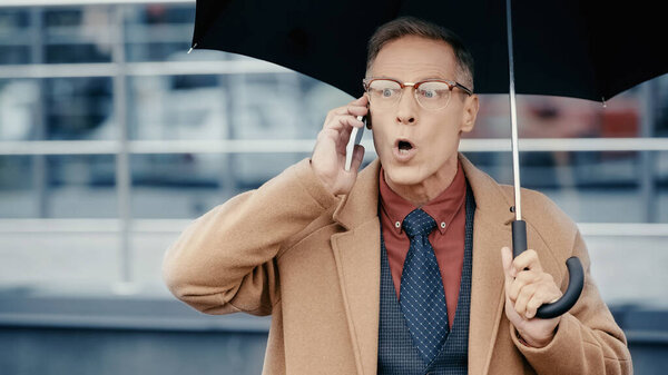 Shocked businessman in coat talking on smartphone and holding umbrella outdoors 