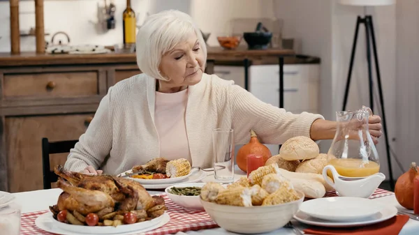 senior woman reaching glass of orange juice near roasted turkey and grilled corn on table