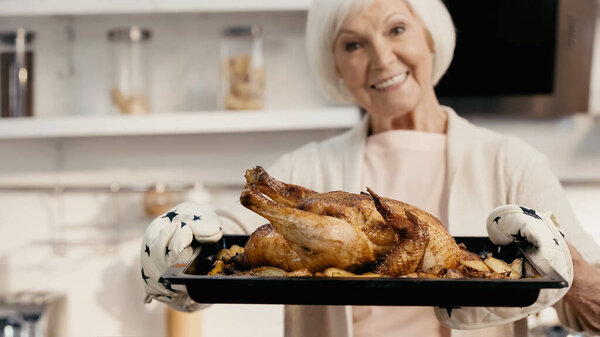 cheerful senior woman holding oven sheet with roasted turkey and potatoes, blurred background