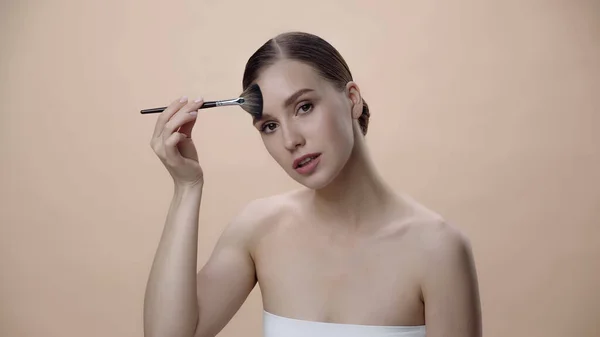 young woman applying face powder while holding cosmetic brush isolated on beige