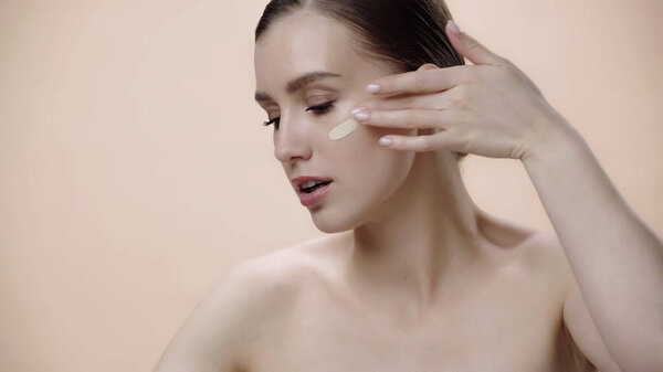 pretty young woman with bare shoulders applying face foundation isolated on beige 