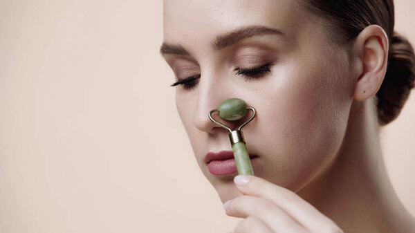 young woman using jade roller on nose isolated on beige 
