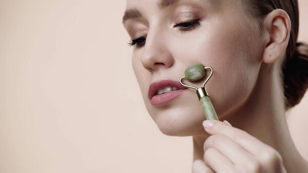 young woman using jade roller on face isolated on beige 