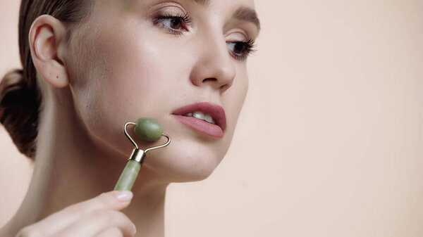 young woman using jade roller while massaging and lifting face isolated on beige 