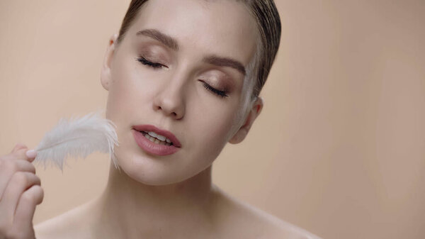 young woman with closed eyes and perfect skin holding white feather isolated on beige 
