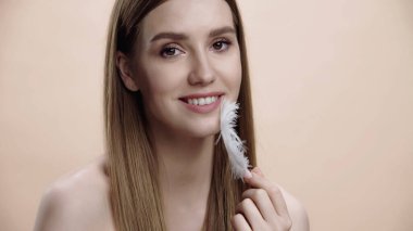 happy young woman with bare shoulders touching face with white feather isolated on beige 