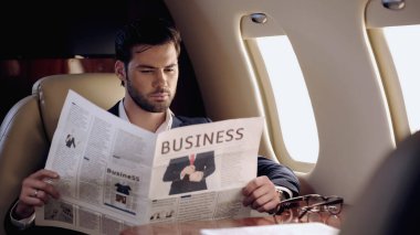 Bearded businessman reading newspaper near eyeglasses in private plane 