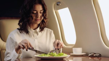 Young businesswoman holding cutlery near salad and coffee in private plane 