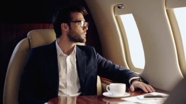 Side view of businessman looking at window near coffee and smartphone in private plane 