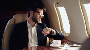 Businessman looking at wristwatch near coffee and cellphone in private plane  clipart