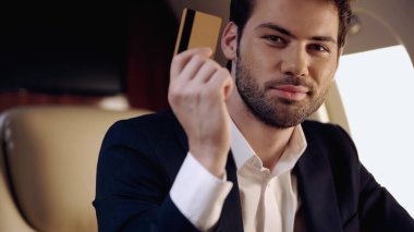 Bearded businessman holding credit card and looking at camera in private plane 
