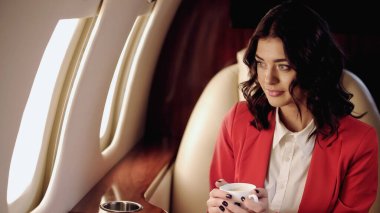 Young businesswoman holding coffee and looking at windows in private jet 