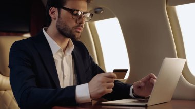 Businessman in eyeglasses holding credit card and laptop in private jet 