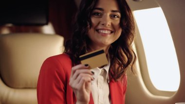Smiling businesswoman holding blurred credit card and looking at camera in private jet 
