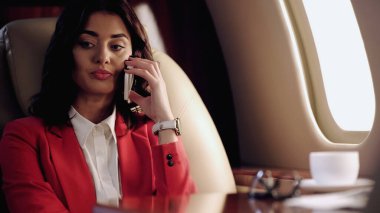 Businesswoman talking on smartphone near blurred cup in private plane 