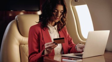 Young businesswoman holding credit card near blurred devices in private plane 