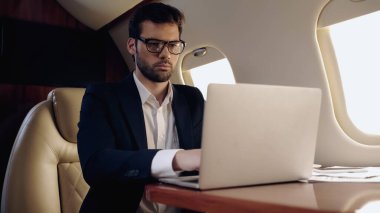 Businessman using laptop near cellphone and papers in private plane 