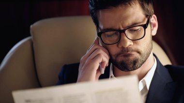 Pensive businessman talking on cellphone while reading blurred newspaper in private plane 