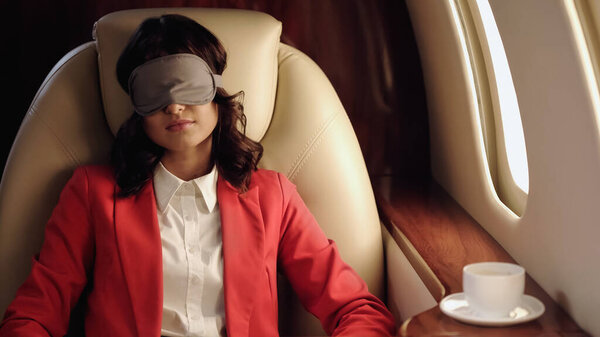 Businesswoman in sleep mask sitting near coffee cup in private plane 