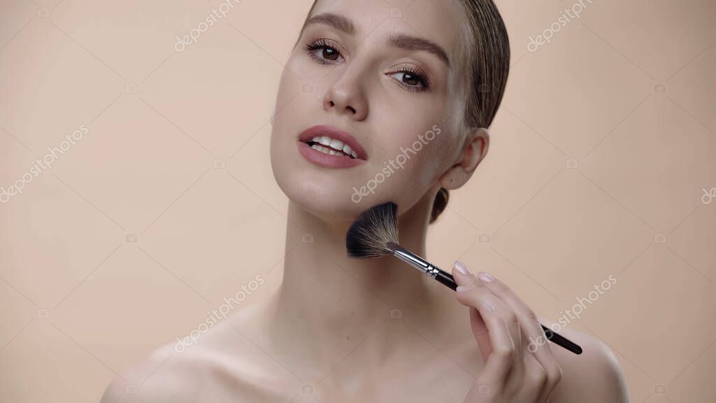 Young woman applying face powder with cosmetic brush isolated on beige