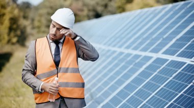 businessman in safety vest rubbing eye and holding bottle with water near solar panels