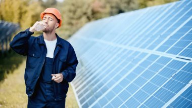 engineer in hardhat and safety vest drinking water near solar batteries 