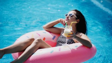 young woman in sunglasses holding glass with cocktail and swimming on inflatable ring in pool