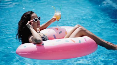 pleased woman adjusting sunglasses while holding glass with cocktail and swimming on inflatable ring in pool