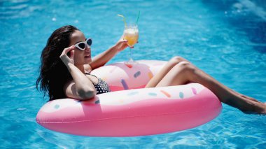 pleased woman adjusting sunglasses while holding glass with cocktail and swimming on inflatable ring in pool clipart