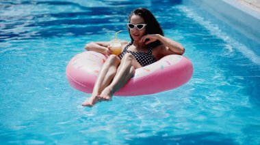 brunette woman in stylish sunglasses holding glass with cocktail and swimming on inflatable ring in pool