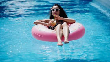 pleased woman in stylish sunglasses drinking cocktail and swimming on inflatable ring in pool