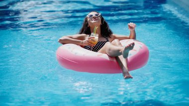 positive woman in stylish sunglasses holding glass with cocktail and swimming on inflatable ring in pool