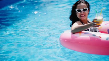cheerful woman in stylish sunglasses holding glass with cocktail and swimming on inflatable ring in pool clipart