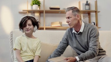 middle aged grandfather holding book near grandson in headphones