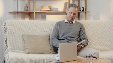 middle aged man reading book near laptop in living room
