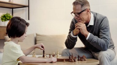 pleased grandfather in glasses and smart grandson playing chess in living room