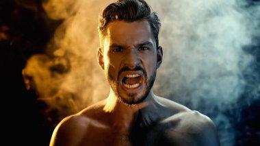 Portrait of irritated sportsman screaming and looking at camera with smoke on black