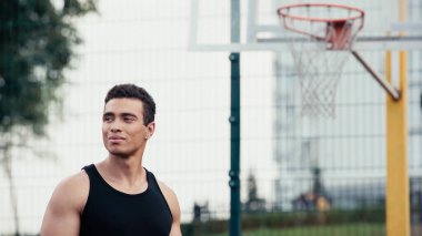 Atletik, çift ırklı bir adam açık havada, bulanık basketbol sahasının yanından uzağa bakıyor.