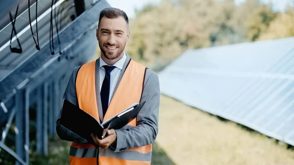 happy businessman in safety vest holding folder near solar panels