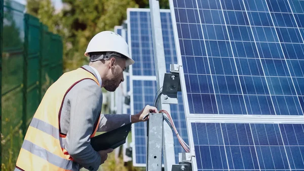businessman in hardhat checking wires and holding folder near solar batteries outside