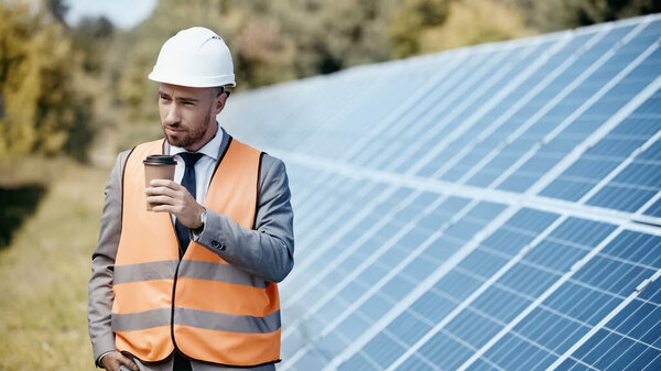businessman in hardhat holding coffee to go near solar batteries outside