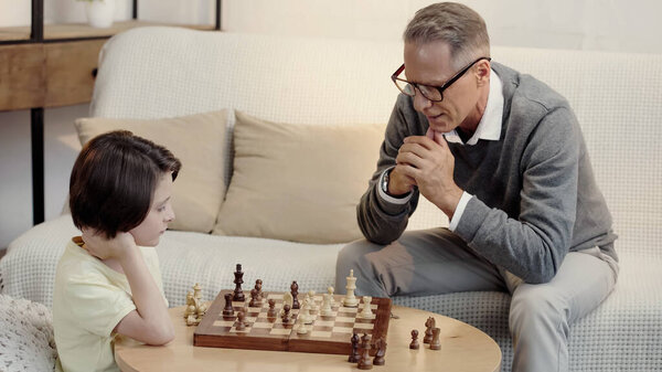 pensive grandfather in glasses and smart grandson playing chess in living room