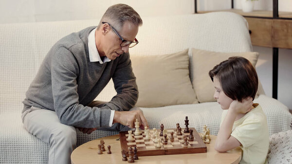 grandfather in glasses and preteen boy playing chess in living room