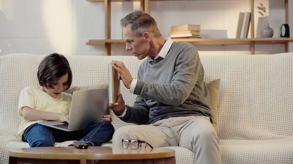 Middle Aged Grandfather Showing Book Grandson Using Laptop Living Room — стоковое фото