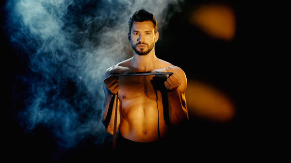 sportsman training with resistance band near smoke behind on black