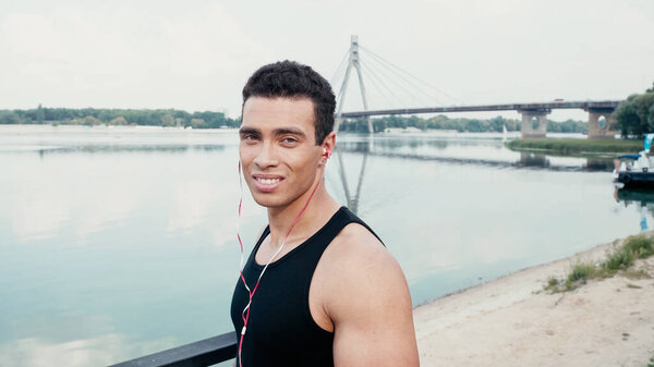 sportive bi-racial man in earphones smiling at camera near river